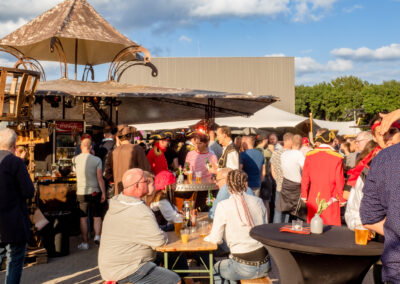 Een buitenevenement in piratenthema met een decoratieve bar onder een uniek ontworpen luifel. Gasten in kostuums en casual kleding genieten van het zonnige weer en de feestelijke sfeer, zittend aan houten tafels of staand in groepen.