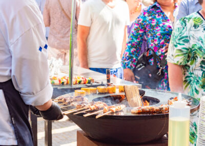 Groepsbarbecue met spiesjes en gegrilde groenten op een open grill, met gasten op de achtergrond.