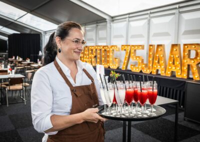 Hostess in een bruine schort serveert een schaal met feestelijke drankjes in een evenementenruimte met verlichte letters '75 jaar.