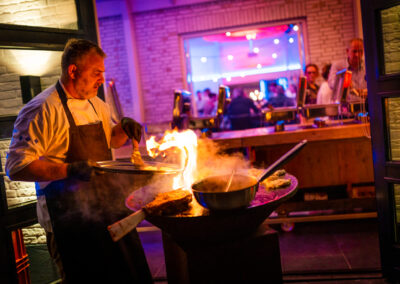 Chef-kok bij een open vuur kookstation, roerend in een pan terwijl vlammen oplichten. De achtergrond toont een sfeervolle locatie met gasten en een kleurrijke ambiance.