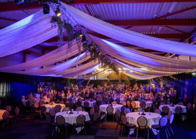 Grote evenementenhal met ronde gedekte tafels en feestverlichting. Het plafond is versierd met witte, golvende doeken, en gasten zitten aan tafels verlicht door kaarsen.