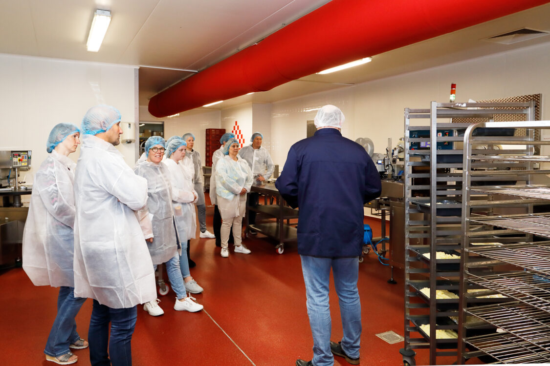 Groep mensen in witte hygiënische overalls en blauwe haarnetjes luistert naar een gids in een productieomgeving. De ruimte heeft een rode vloer en moderne apparatuur, waaronder rekken en machines, geschikt voor voedselverwerking. De gids staat met de rug naar de camera en legt het proces uit.