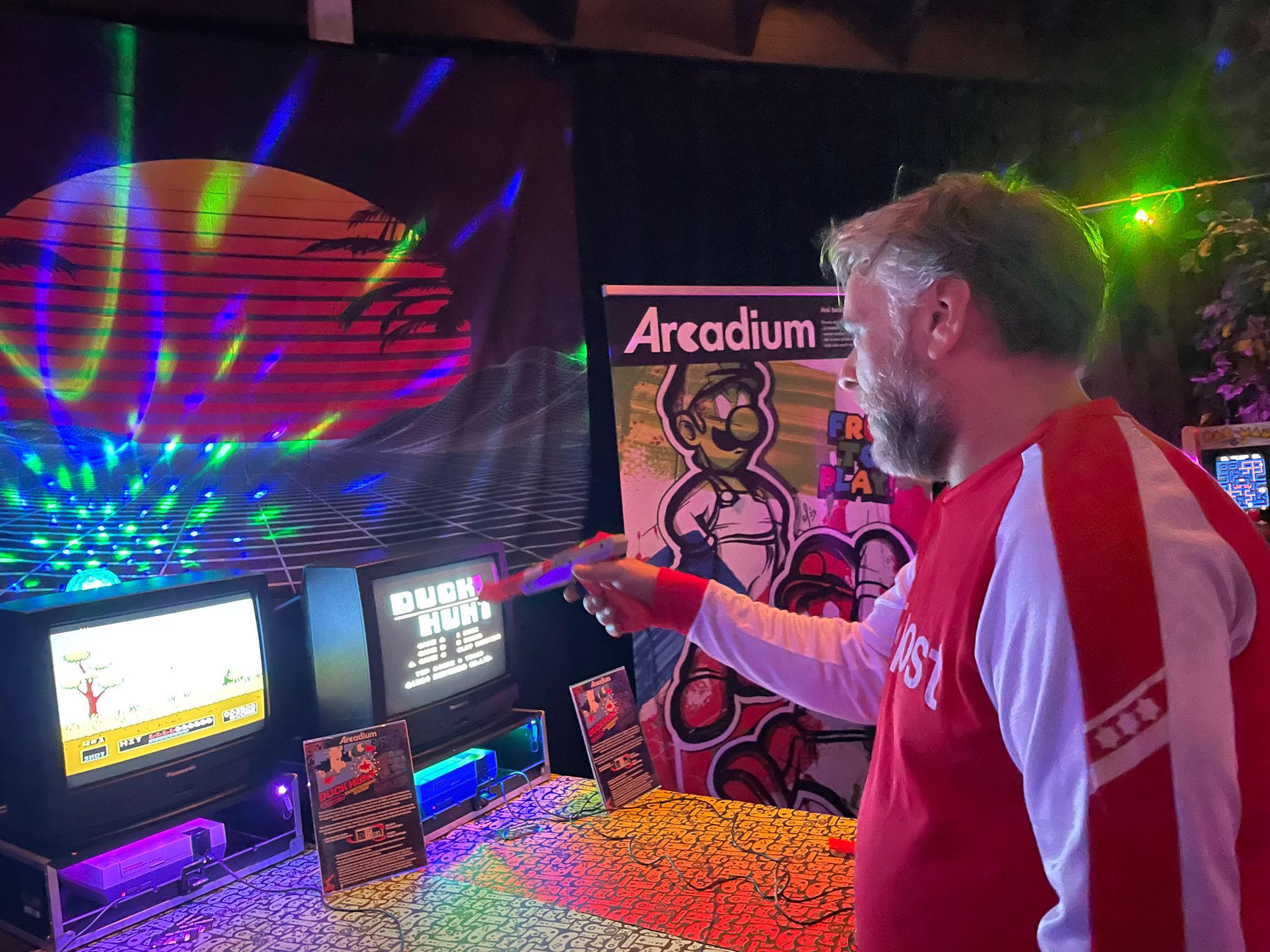 Man in retro kleding speelt Duck Hunt op een klassieke spelconsole, met kleurrijke neonverlichting en een retro Arcadium-thema op de achtergrond.