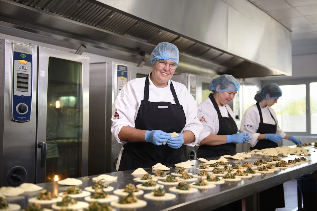 Een groep koks in de productiekeuken die voedsel bereiden, gekleed in witte kokskleding, zwarte schorten en blauwe haarnetjes en handschoenen. Ze zijn bezig met het vullen van deeg met een mengsel van groenten