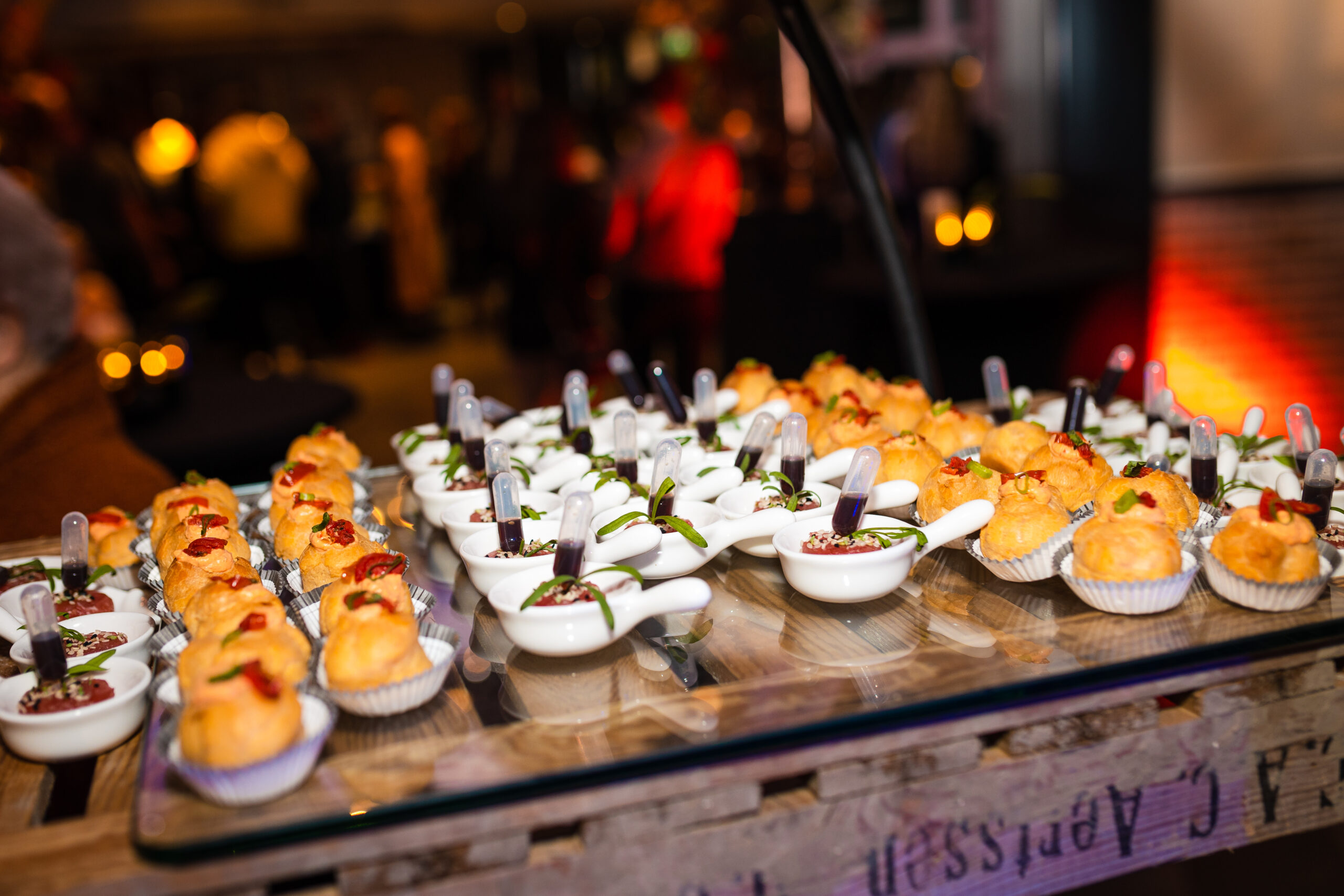 Een tafel vol met verfijnde hapjes, waaronder gevulde deegwaren en kleine witte schaaltjes met culinaire creaties, versierd met kleine pipetten met saus. De achtergrond is vaag, met mensen die deelnemen aan een evenement in een sfeervolle omgeving met warme verlichting.