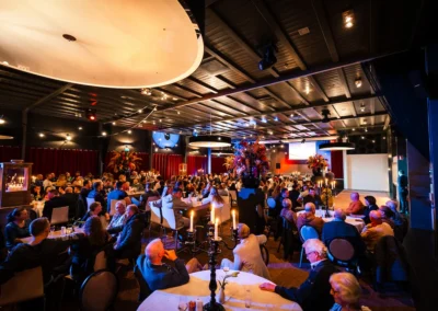 Gasten genieten van een sfeervol evenement in De Koningshof, georganiseerd door Willem de Boer. De zaal is prachtig verlicht en sfeervol aangekleed met kaarsen en bloemen.