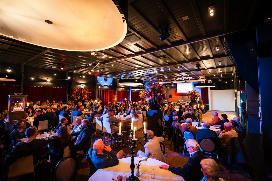 Gasten genieten van een sfeervol evenement in De Koningshof, georganiseerd door Willem de Boer. De zaal is prachtig verlicht en sfeervol aangekleed met kaarsen en bloemen.