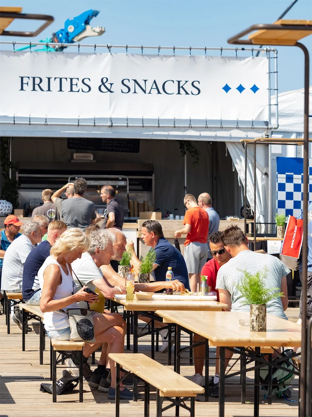 Mensen genieten van hun maaltijden aan lange tafels bij een 'Frites & Snacks'-kraam tijdens een de technische kontact dagen.