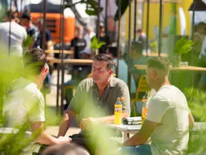 Drie mannen zitten buiten aan een tafel en drinken frisdrank tijdens een evenement, met andere mensen en stands zichtbaar op de achtergrond. De scène is zonnig en omringd door groene planten.