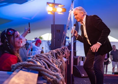 Een artiest entertaint het publiek tijdens een avondoptreden, terwijl een vrouw in piratenkostuum naar hem luistert