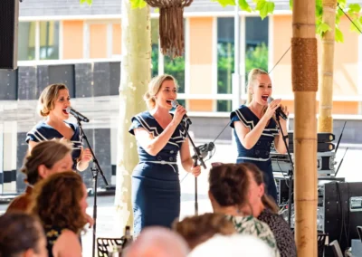 Een trio zangeressen treedt op tijdens een zomers evenement, gekleed in marineblauwe outfits, terwijl het publiek geniet van de muziek