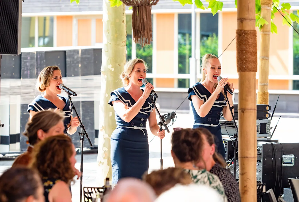 Een trio zangeressen treedt op tijdens een zomers evenement, gekleed in marineblauwe outfits, terwijl het publiek geniet van de muziek