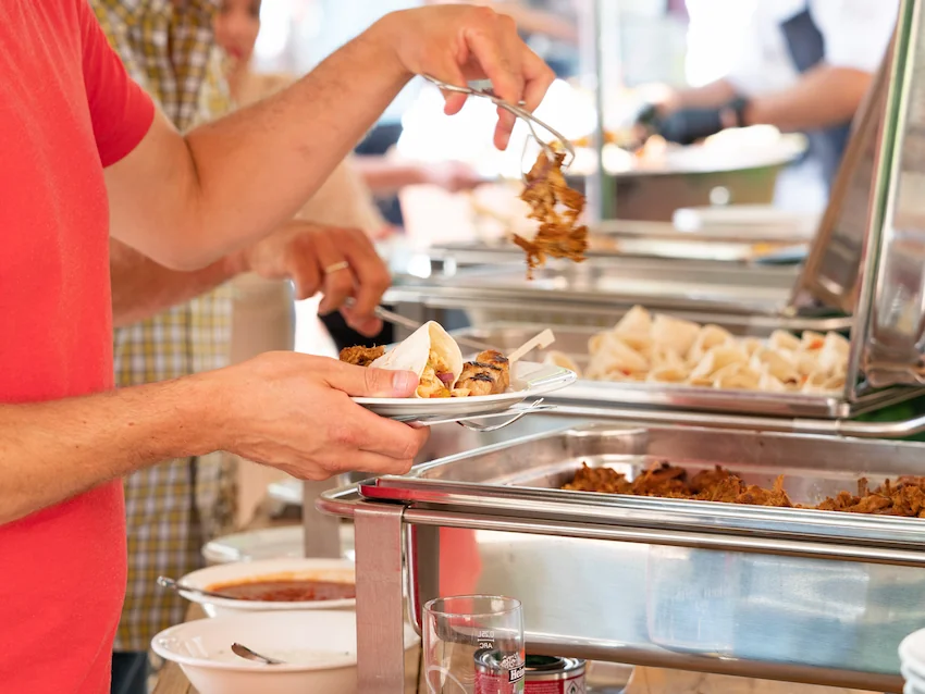 Een persoon schept eten op een bord bij een buffet, met verschillende warme gerechten en sauzen.