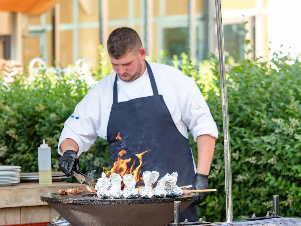 Een chef-kok grilt gerechten op een buitenbarbecue tijdens een evenement, met vurige vlammen en een groene achtergrond