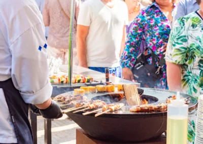 Een kok grilt spiesjes en maiskolven op een buitenbarbecue, terwijl bezoekers in de rij staan tijdens een zomers evenement