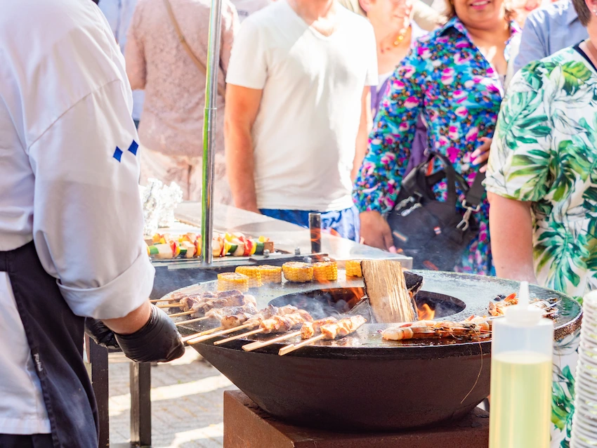 Een kok grilt spiesjes en maiskolven op een buitenbarbecue, terwijl bezoekers in de rij staan tijdens een zomers evenement