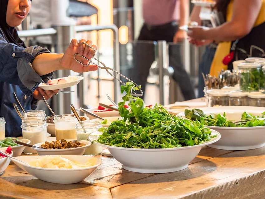 Een persoon schept verse groenten op tijdens een buffet, omringd door verschillende salades en dressings op een houten tafel