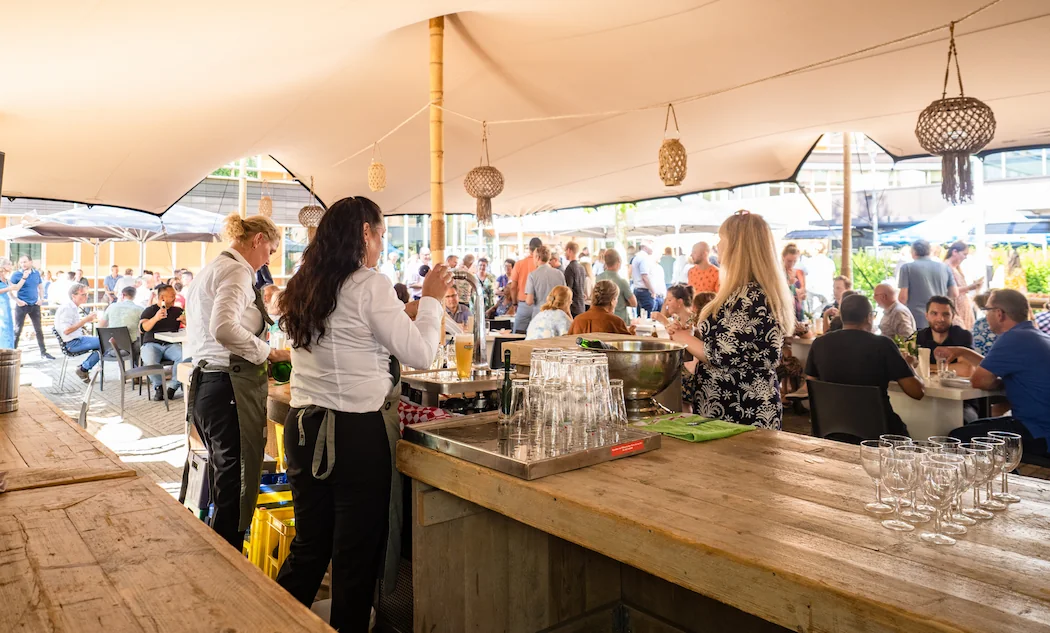 Serveerders bereiden drankjes voor gasten tijdens een druk bezocht buitenfeest onder een grote tent