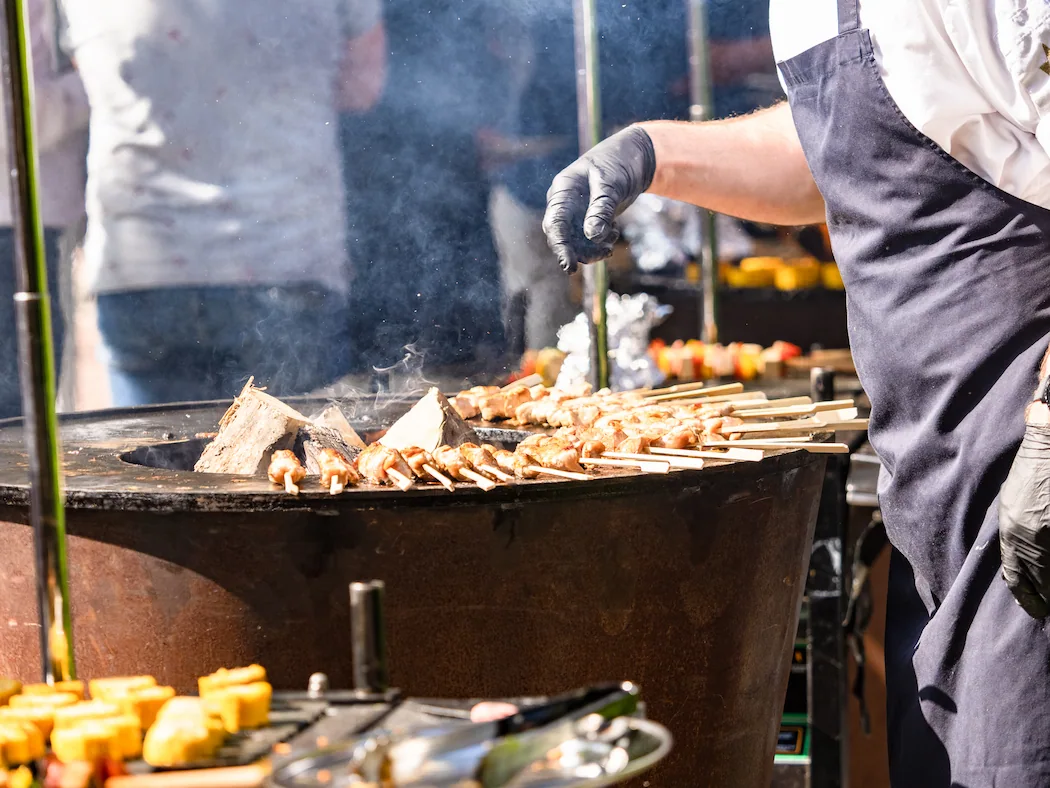 Een chef-kok grilt kipsatés en andere gerechten op een buiten barbecue, met rook opstijgend van de grill.