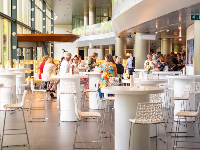 Gasten genieten van een bijeenkomst in een moderne ruimte met grote ramen, ronde tafels en comfortabele stoelen