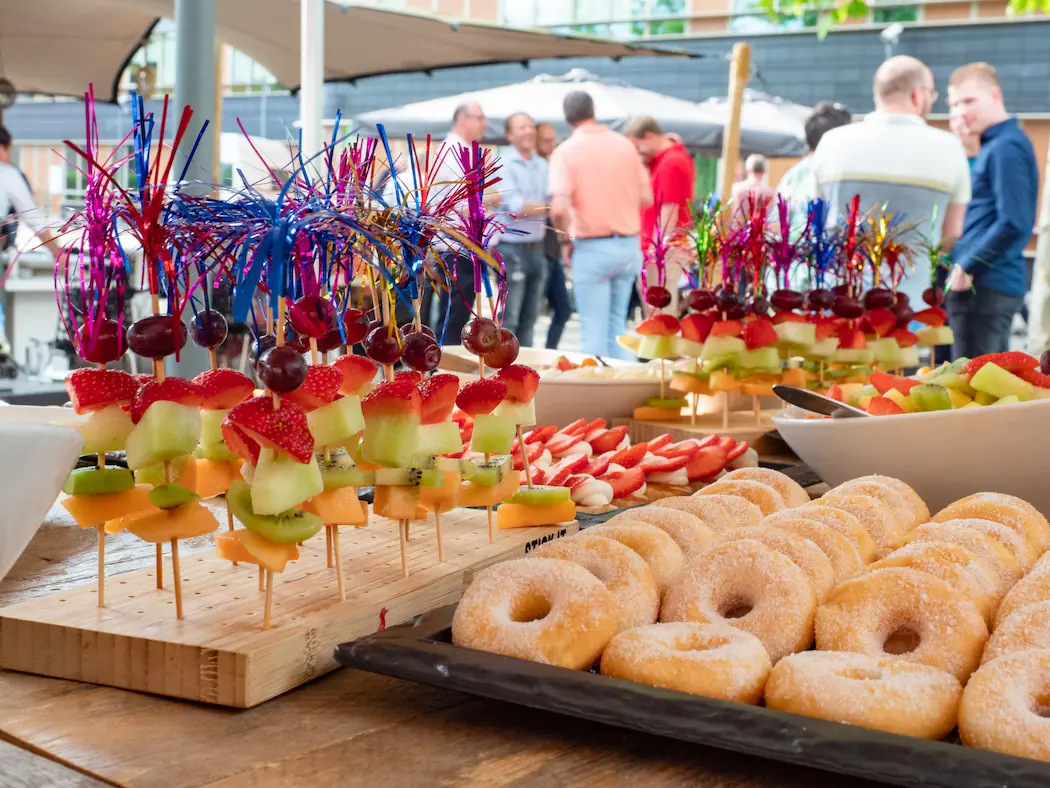 Feestelijke fruitspiesjes en donuts op een tafel tijdens een zomerse bijeenkomst, met mensen op de achtergrond die genieten van het evenement