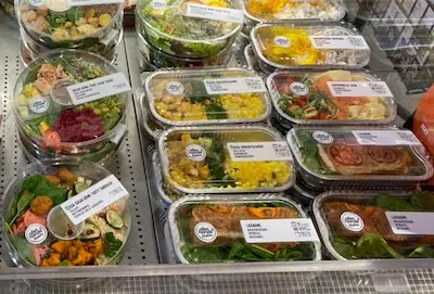 Een display met verschillende voorverpakte salades en maaltijden in een gekoelde vitrine, met etiketten die de inhoud en prijzen aangeven. De verpakkingen variëren in vorm, waaronder ronde en rechthoekige bakjes, en bevatten diverse ingrediënten zoals groenten, vlees en granen.