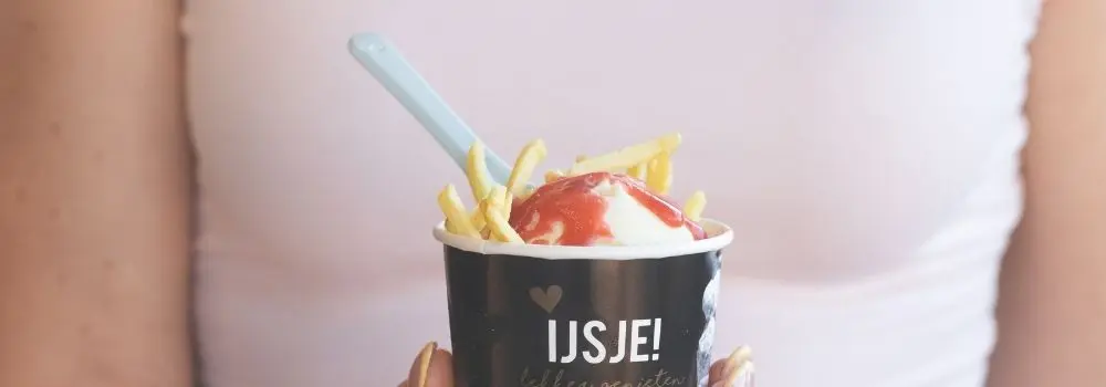 Willem IJs mayonaise ijs in een zwart bakje met frietjes en aardbeiensaus, vastgehouden in de hand tegen een lichte achtergrond.